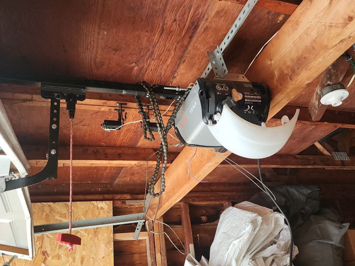 Chain-drive opener fixed to the exposed rafters of an unfinished garage ceiling, rail, chain and manual release cord visible